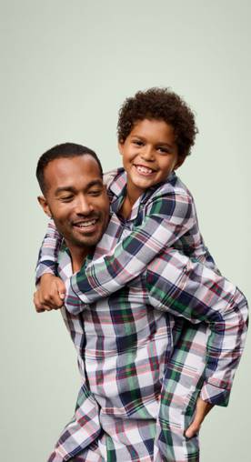 A father giving his smiling child a piggyback ride, both wearing matching plaid pajamas against a light green background.