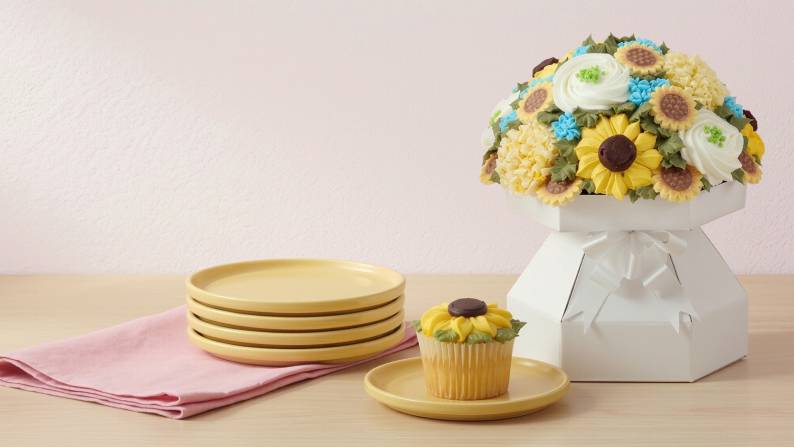 A stack of plates next to a sunflower inspired cupcake on a plate and a bouquet of cupcakes decorated to look like flowers on a wooden countertop.