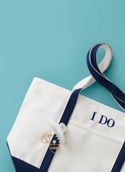 A blue and white tote bag with I do embroidered on it, a pair of heart-shaped earrings and a white hair clip are pictured on a blue backdrop.