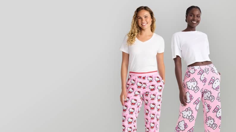 Two women wearing white tops and pink Hello Kitty pajama bottoms while standing in front of a gray background.