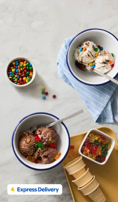 Two bowls of chocolate and vanilla ice cream topped with candy, next to waffle cones and candy toppings.