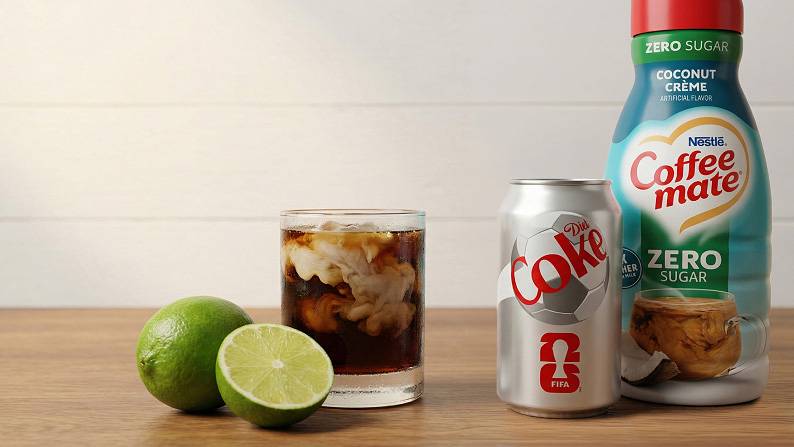 Fresh limes, a glass with cola and creamer, a can of Diet Coke, and a bottle of Coffee mate zero sugar coconut creme creamer on a wood counter.
