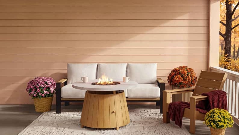 Round fire pit table with two mugs between sofa and wooden chairs on covered patio with fall flowers and rug.