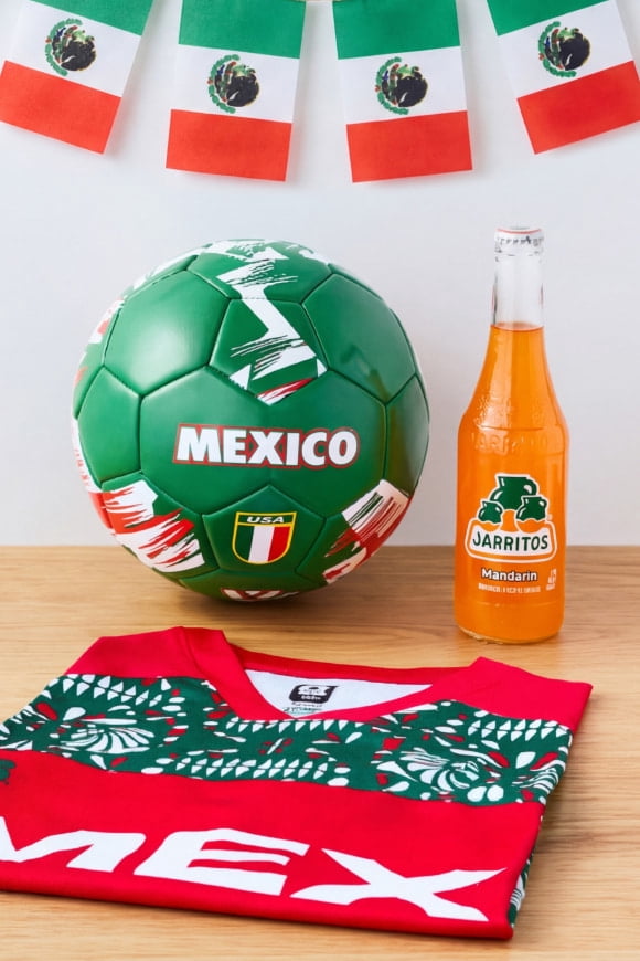 A variety of Team Mexico memorabilia is on a wooden counter—a jersey, a soccer ball, a bottle of Jarritos and a flag banner.