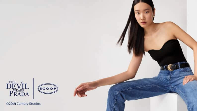 Model sits in studio wearing black strapless top and blue jeans with black belt and gold buckle looking at camera.