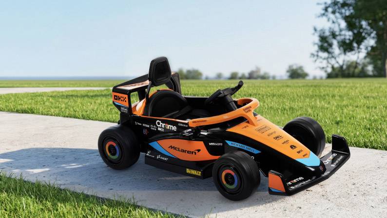 A powered ride-on with orange and blue details on a concrete path in an outdoor setting with grass and trees.