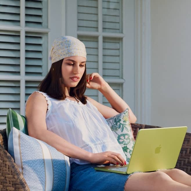 A woman in a bandana types on a lime green laptop while sitting outdoors on a wicker sofa with blue & green pillows.