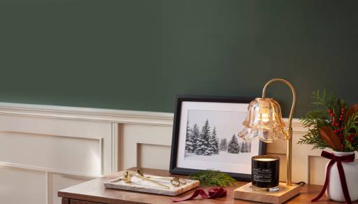 Side table with framed snowy tree art, lit candle lamp, greenery, red berries and ribbon for a cozy holiday display.