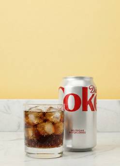 A glass of Diet Coke on ice next to the can on a marble countertop with a yellow background.