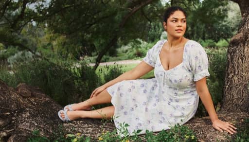 A woman in a white floral dress and blue sandals sits on a tree trunk in a lush green park, surrounded by grass and trees.