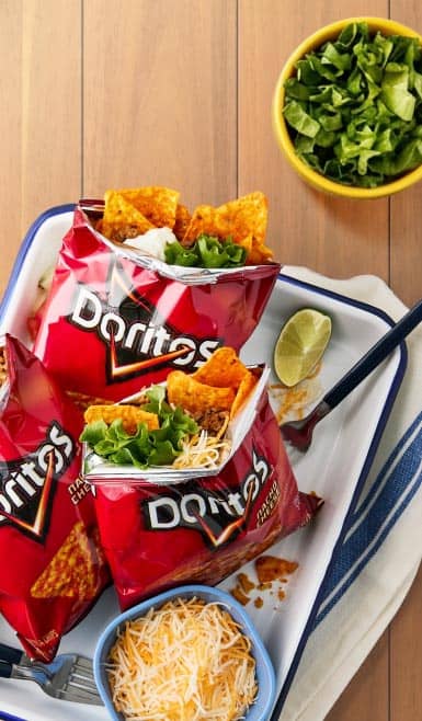 Three walking tacos in bags of Doritos alongside bowls of toppings on a table.