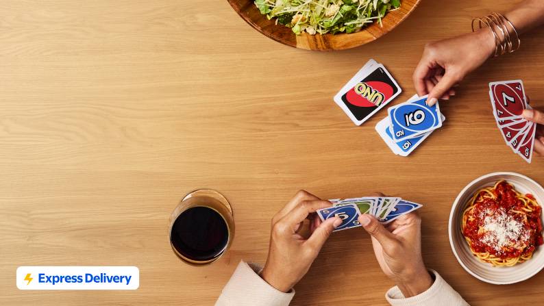 Express Delivery badge with dinner, drinks, and a card game for two on a table, alongside pasta, bread, and wine.