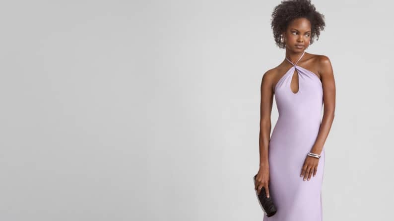 A woman is wearing a lavender dress with a key-hole cutout and is holding a black clutch.