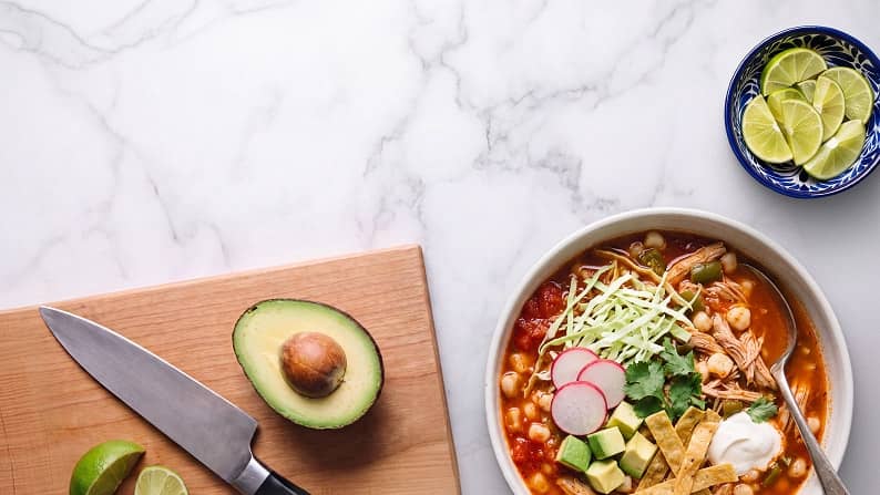 Bowl of soup with shredded meat, cabbage, radish, avocado and tortilla strips beside cutting board with knife, avocado and lime wedges.