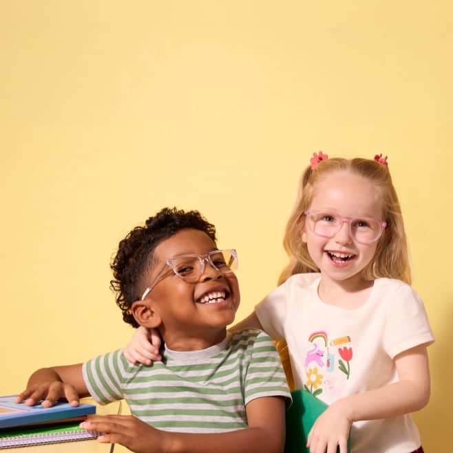 Two children wearing glasses and smiling while one sits at a desk.