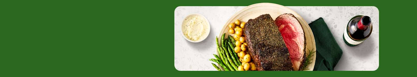 Prime rib, roasted baby potatoes, and asparagus on a platter next to a bowl of horseradish sauce and a bottle of red wine on a white granite counter.