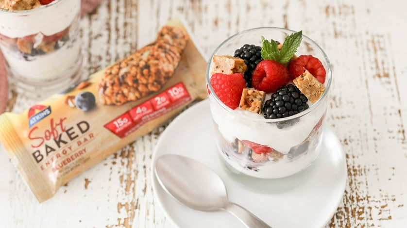 A fruit and yoghurt parfait in a glass on a rustic looking table next to an Atkins soft baked bar