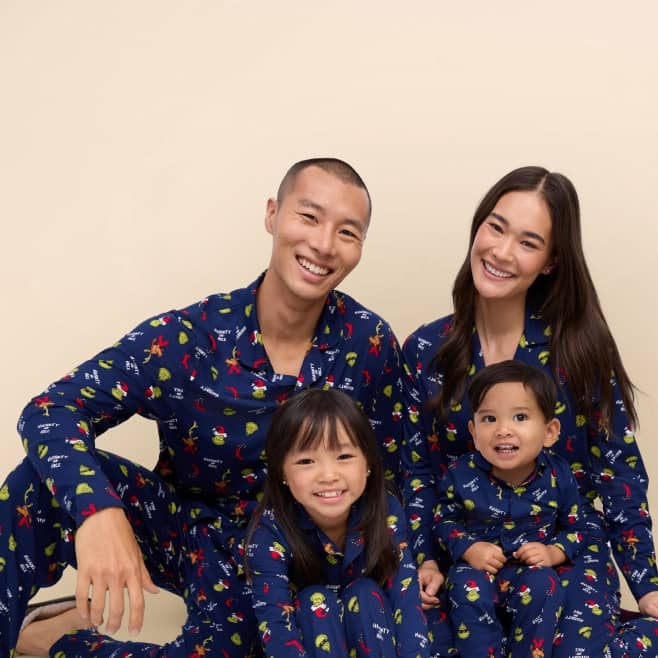 Two adults and two children sit together wearing matching navy pajamas with colorful fruit and holiday motifs.