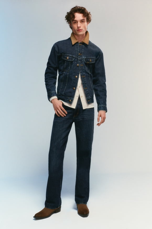 Young adult man wearing a dark denim jacket with a tan corduroy collar, cream button-up, dark jeans and brown suede boots.