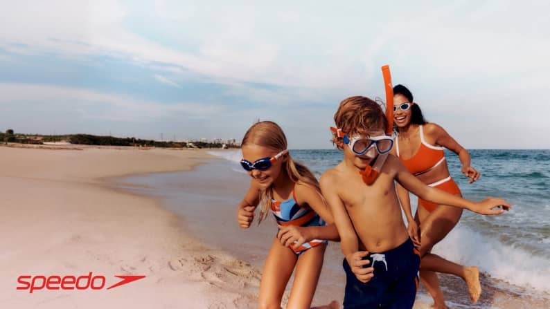 Speedo logo. A young boy, a young girl, and a woman with Speedo swimming accessories play in the ocean wearing Speedo swimsuits.