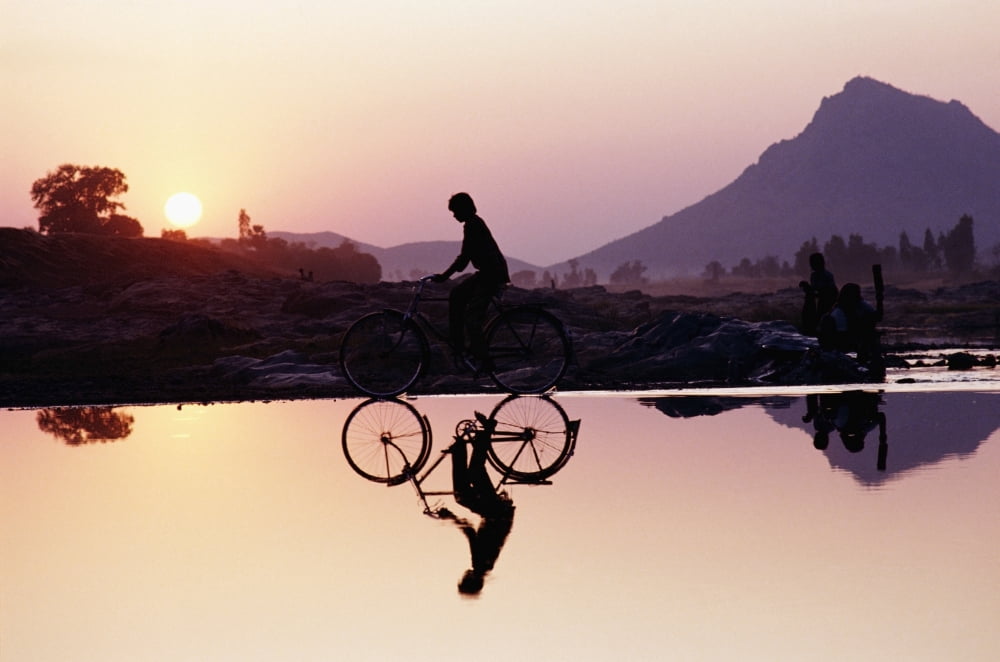 Bicyclist Crossing Shallow River At Sunset With Women In Background Doing Washing Canvas Art - Paul Quayle Design Pics (17 x 11)