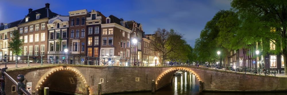 Buildings and arched bridges at the intersection of Herengracht and Reguliersgracht at night Amsterdam North Holland Netherlands Canvas Art - Panoramic Images (36 x 12)