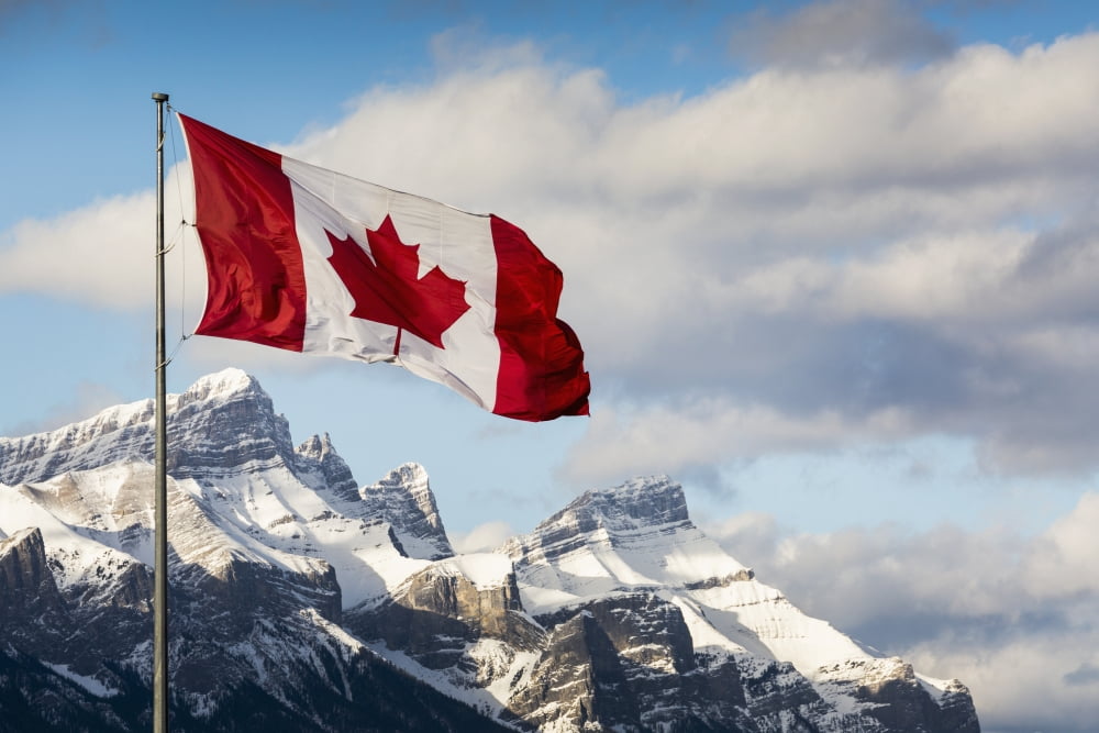 Canadian flag blowing in the wind on a flag pole with snow covered mountain range in the background with blue sky and clouds Canmore Alberta Canada Canvas Art - Michael Interisano Design Pics (38 x 2