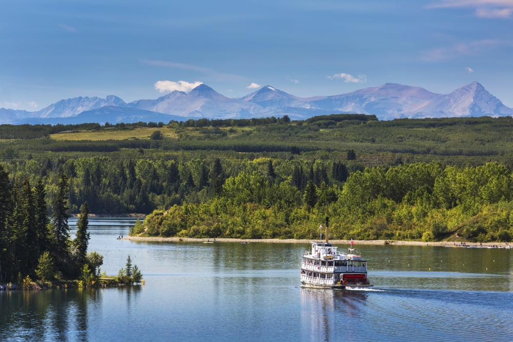 Paddlewheel boat on lake with tree lined shoreline hills and mountains in the background and blue sky and clouds Calgary Alberta Canada Canvas Art - Michael Interisano  Design Pics (38 x 24)