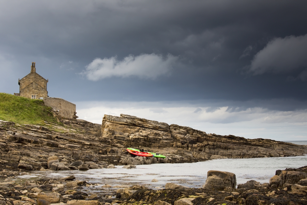 Red And Green Kayaks Sitting On The Rocky Shore With A House At The Waters Edge Northumberland England Canvas Art - John Short Design Pics (19 x 12)