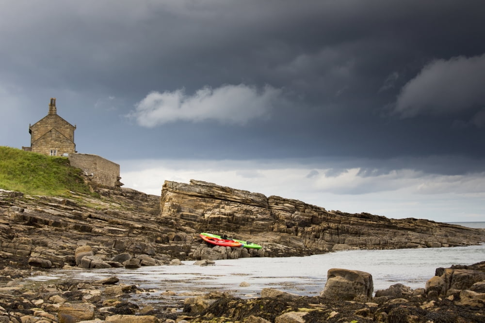 Red And Green Kayaks Sitting On The Rocky Shore With A House At The Waters Edge Northumberland England Canvas Art - John Short Design Pics (38 x 24)