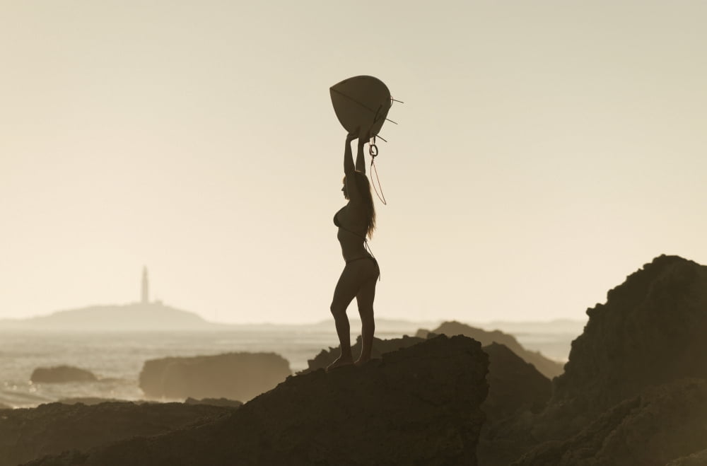 Silhouette of a female surfer holding her surfboard up above her head while standing on a rock at the coast andalusia spain Canvas Art - Ben Welsh Design Pics (19 x 12)