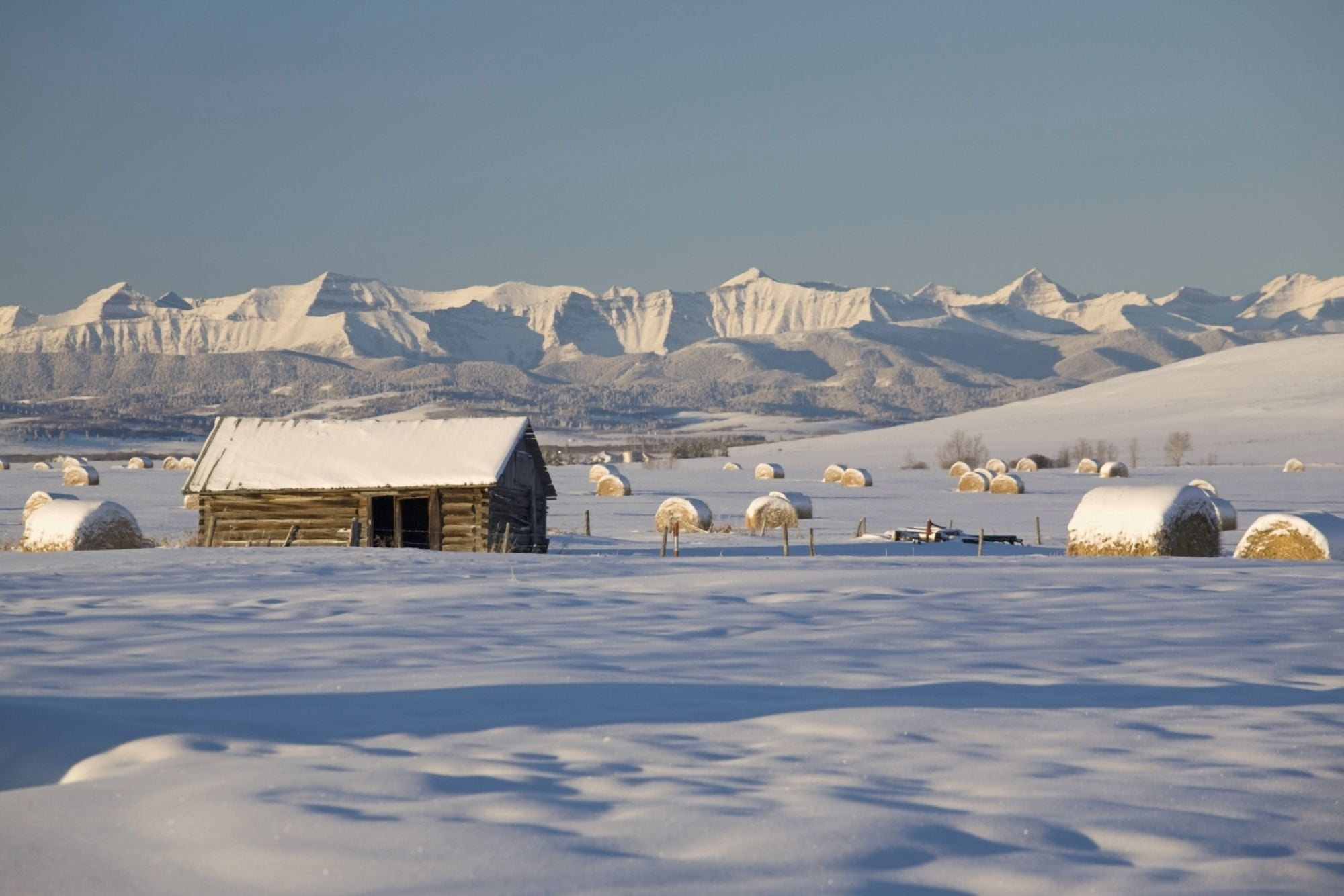 Snow Covered Log Cabin And Hay Bales In A Field With Snow Covered Mountains Okotoks Alberta Canada Canvas Art - Michael Interisano Design Pics (19 x 12)