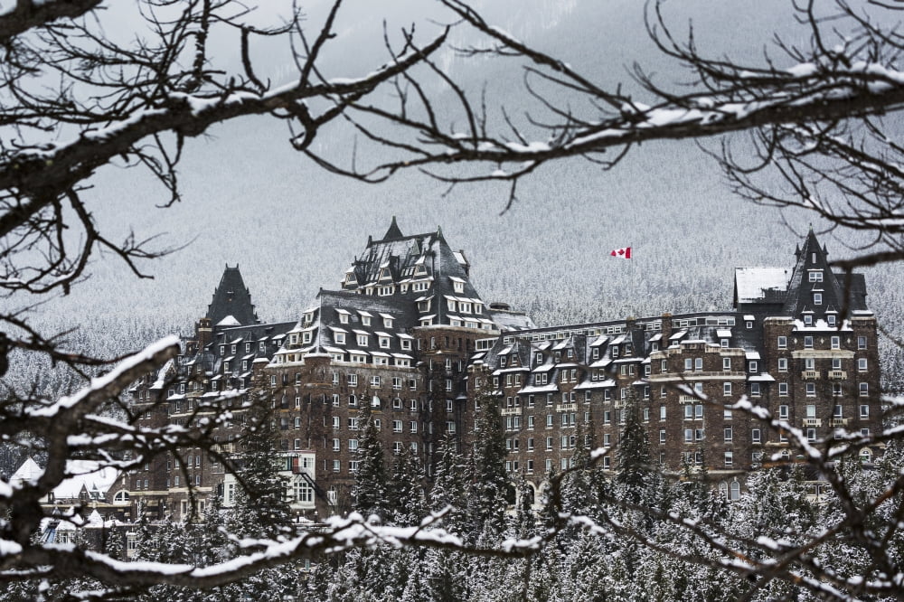 Snow covered Banff Springs Hotel framed by tree limbs with snow covered trees on mountain hillside in the background Banff Alberta Canada Canvas Art - Michael Interisano Design Pics (38 x 24)