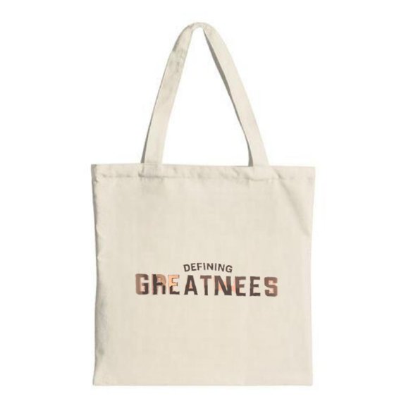 Stylish canvas tote bag with two handles featuring bold " DEFINING GREATESTES " text in metallic font perfect for shopping and travel