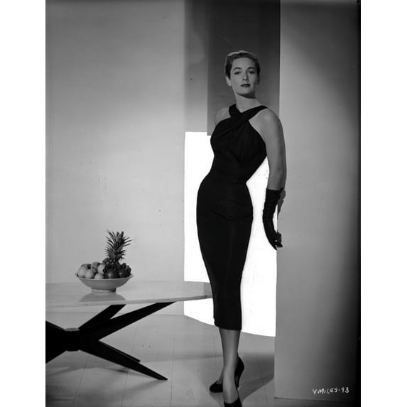 Vera Miles wearing a black, cocktail length, crossed-halter dress , black pumps and black gloves, standing next to a low-sitting table Photo Print (24 x 30)