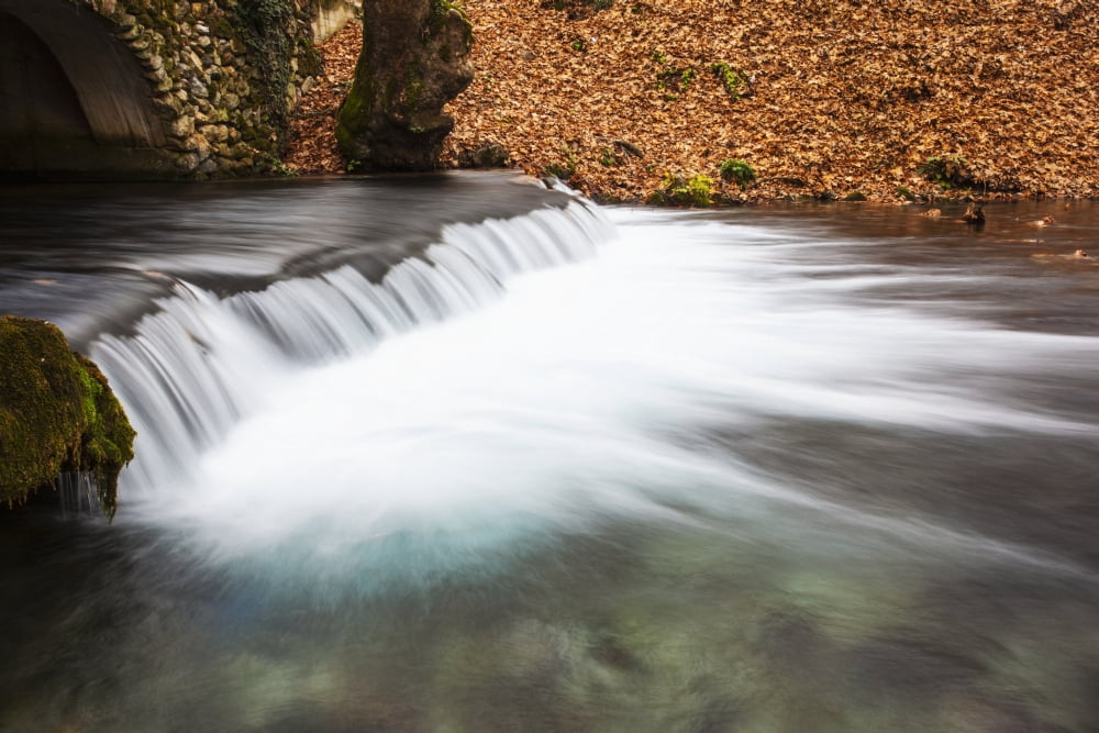 Water cascading over a ledge in a river with the riverbank covered with autumn coloured fallen leaves Naoussa Greece Canvas Art - Reynold Mainse Design Pics (38 x 24)