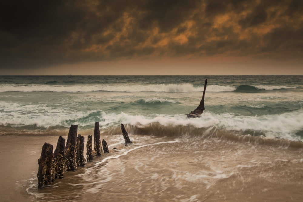 Wooden posts on a beach with a boat being tossed in the water and waves crashing into the sand under a cloudy sky Queensland Australia Canvas Art - John Short Design Pics (19 x 12)