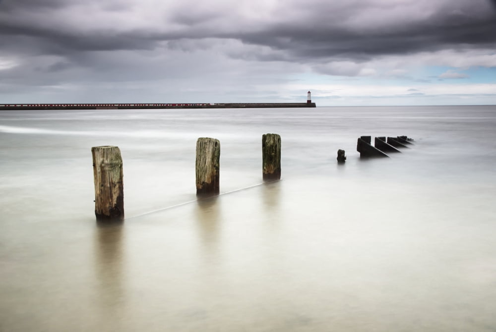 Wooden posts in the ocean with a lighthouse at the end of a pier Canvas Art - John Short Design Pics (18 x 12)