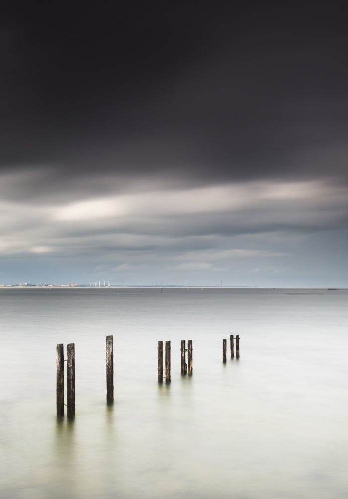 Wooden posts in a row in the shallow water along the coast with a view of a city in the distance under dark storm cloudsSt marys bay northumberland england Canvas Art - John Short  Design Pics (12 x 1