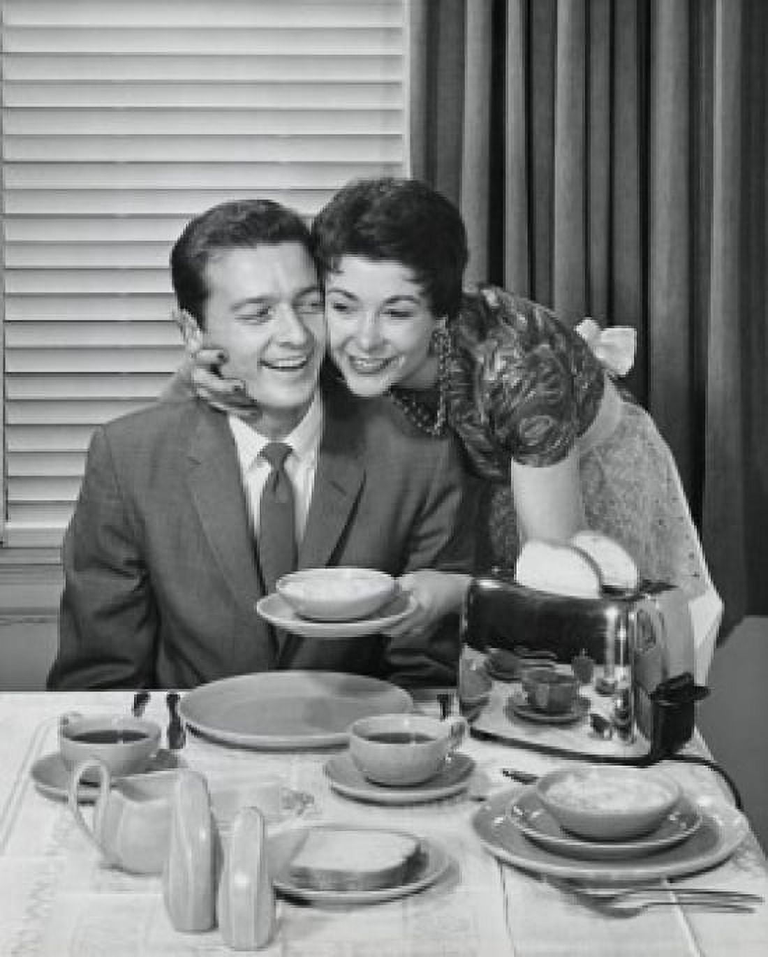 Young woman holding a bowl and embracing a young man sitting at the dining table Canvas Art - (24 x 36)