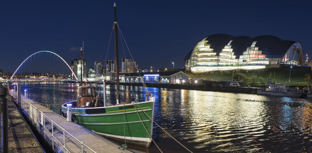 A green fishing boat moored along the edge of a river at nighttime with an illuminated Sage Gateshead in the background Newcastle Tyne and Wear England Canvas Art - John Short Design Pics (44 x 22)