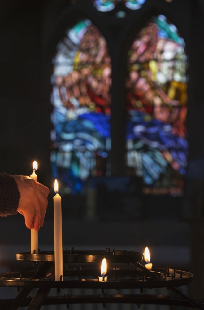 A mans hand lights a candle in a church with colourful stained glass window in the background Northumberland England Canvas Art - John Short Design Pics (12 x 19)